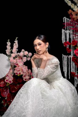 Endonezyalı gelin bembeyaz süslenmiş bir elbiseyle elleri çenesinin altında oturuyor, kusursuz bir gelin makyajı yapıyor, zarif mücevherler takıyor ve etrafı romantik çiçeklerle çevrili..