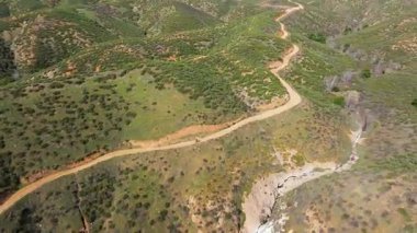 Çimenli bir yamaçtan geçen toprak bir yol. Yol kayalık ve engebeli. Arka planda ağaçlar var.