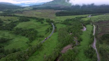 İçinden nehir akan yeşil bir tarla. Nehir ağaçlarla çevrilidir. Gökyüzü bulutlu.
