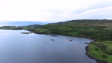 Arkasında yeşil bir tepe olan bir su kütlesi. Su sakin ve berrak. Arka planda ağaçlar var.