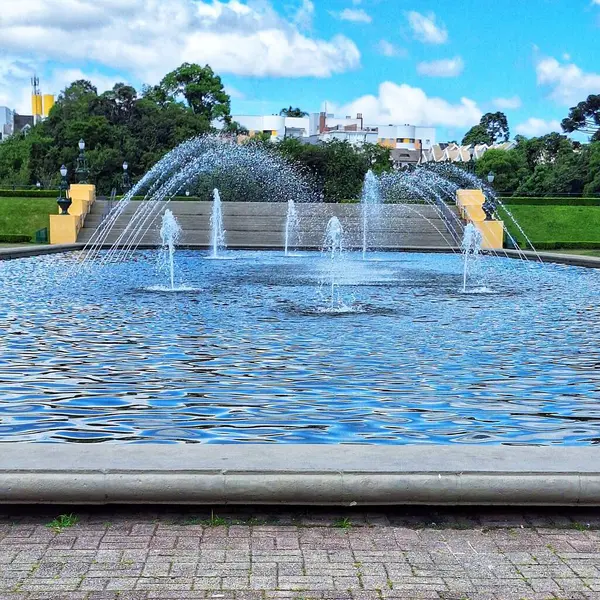 Şehir parkında büyüleyici bir çeşme, su fışkıran fıskiyeler, güzel, güneşli bir günde güneş ışığını yansıtıyor. Arka planda yeşil ağaçlar ve beyaz bulutlarla benekli mavi bir gökyüzü ile ortam rahatlatıcı. Çevrenin sakinliği