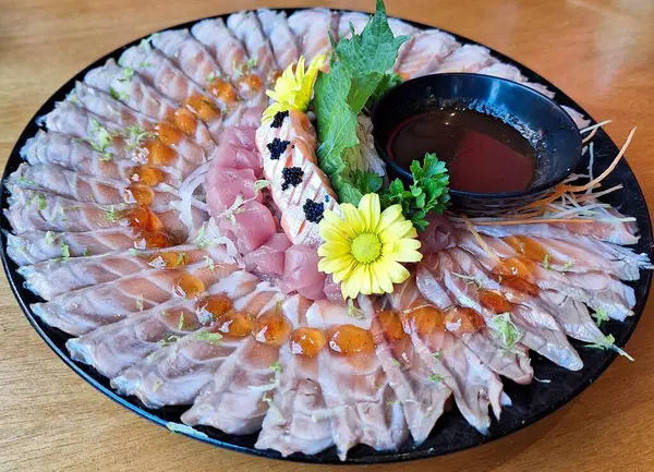 Zarif bir Japon saşimi tabağının çarpıcı görüntüsü ince dilimler halinde sanatsal bir şekilde düzenlenmiş, sarı çiçekler ve yanında koyu bir sos ile süslenmiş, tazelik ve inceliği çağrıştırıyor..