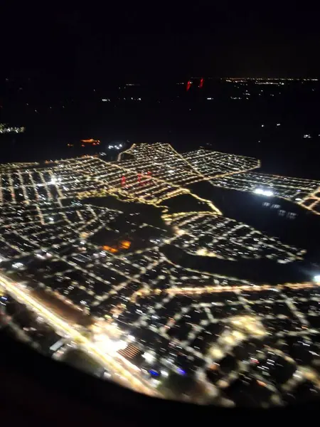 Aydınlık sokaklar ve lambaların altında görünen yerleşim alanlarıyla aydınlık bir şehrin etkileyici bir gece hava görüntüsü. Gölde yansıtılan, güzel, sakin bir manzara yaratır. Renkli trafik ve bina ışıkları karanlığa karşı duruyor