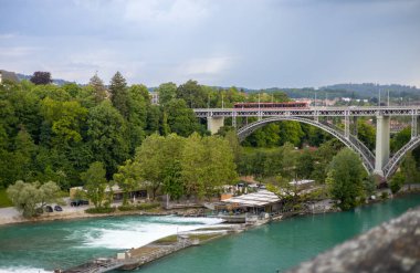 Bern, İsviçre, 20 Haziran 2024. Kızıl bir tramvay, İsviçre 'nin Bern kentindeki turkuaz Aare Nehri üzerinden tarihi Kirchenfeld Köprüsü' nü geçer. Zarif metal kemer köprüsü yemyeşil ile çevrilidir.