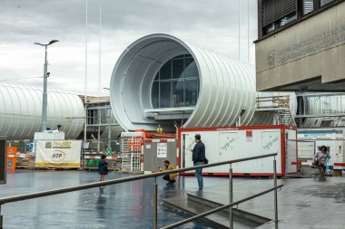 Ziyaretçiler, CERN 'in modern dairesel binalarının önünden bulutlu gökyüzünün altında yürüyorlar.. 