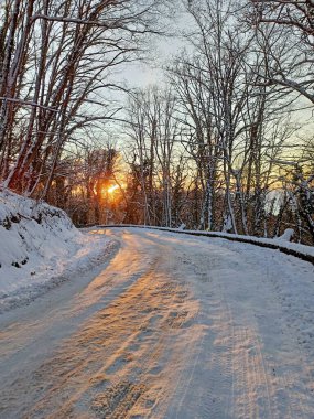 Karla kaplı bir yol içeren sakin bir kış manzarası, batan güneşin sıcak renkleriyle aydınlatılan, don kaplı ağaçlardan oluşan bir orman boyunca kıvrılıyor.. 