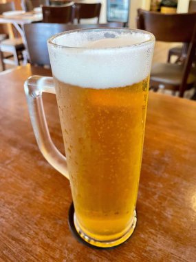 Soğuk birayla dolu ferahlatıcı bir cam bardak, arka planda sandalyeleri olan rahat bir bar veya restoran ortamındaki ahşap bir masaya yerleştirilmiş bir köpük kafası sergiliyor..