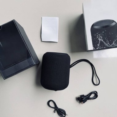 Beyaz bir yüzey üzerinde kayış, USB kablosu ve ambalaj kutusu olan siyah taşınabilir bir bluetooth hoparlörü..