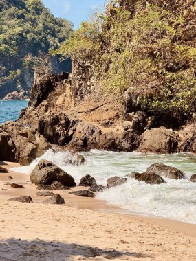 Dalgaların kıyıya vurduğu kayalık bir Teluk Ijo plajının manzarası. Berrak mavi gökyüzünün altında yemyeşil ve kayalıklarla çevrili..