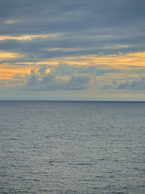 Gün batımında bulutlu bir gökyüzünün altında, yumuşak dalgaların gökyüzünün renklerini yansıttığı sakin bir okyanus manzarası. Payangan Plajı Jemonu