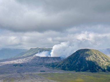 Bereketli yeşil tepeler ve bulutlu gökyüzü ile çevrili bir kraterden yükselen dumanla volkanik bir manzaranın manzarası. Bromo Malang