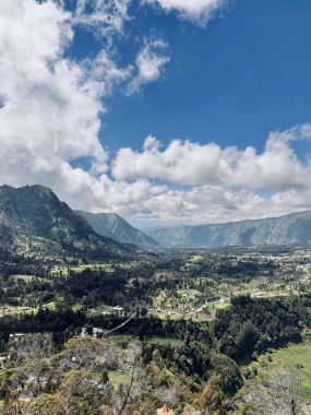 Parçalı bulutlu bir gökyüzünün altındaki dağlarla çevrili yemyeşil bir vadinin panoramik görüntüsü. Manzarada ağaçlar, tarlalar ve uzak tepelerden oluşan sakin bir manzara yer alıyor. Seruni Noktası Bromo