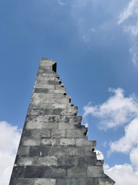 Uzun, açısal taş bir yapı dağınık bulutlu mavi bir gökyüzüne doğru uzanıyor. Yapının eşsiz bir basamak tasarımı var, arkaplana çarpıcı bir siluet oluşturuyor. Seruni Noktası Bromo