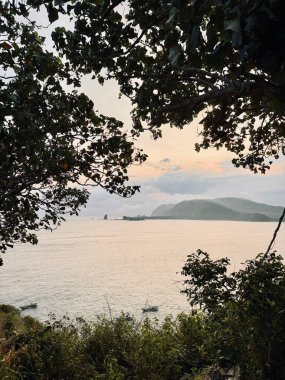 Gün batımında ağaç dalları tarafından çerçevelenmiş sakin bir kıyı manzarası. Sakin deniz, uzak tepeleri ve ufukta küçük bir adası olan gökyüzünün yumuşak renklerini yansıtır. Payangan Beach Jember, Endonezya