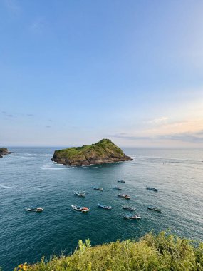 Scenic view of a small island surrounded by the ocean with several boats in the water, under a clear blue sky. Payangan Beach Jember, Indonesia clipart