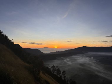 Dağlık bir arazide nefes kesici bir gün doğumu, vadilerden süzülen sis ve geceden güne değişen berrak bir gökyüzü. Bromo Tengger Semeru Ulusal Parkı, Malang, Endonezya