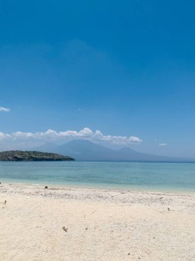 Menjangan Adası, Batı Bali, kristal berrak mavi suları ve el değmemiş beyaz kumlarıyla. Uzaktaki üç görkemli dağ panoramayı çevreleyerek sakin bir doğal atmosfer yaratıyor..