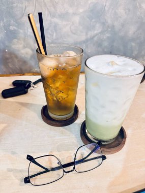 Bir masada iki bardak içecek, biri buzlu çay, diğeri de kremalı yeşil içecek. Masanın üzerinde bir çift gözlük duruyor, anahtarların yanında..