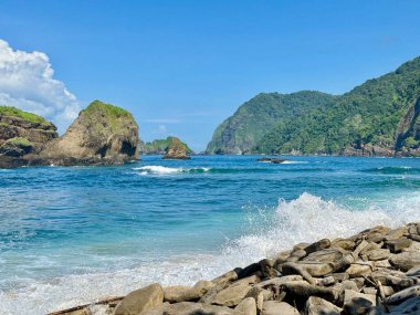 Mavi okyanus dalgaları kayaların üzerinde kırılıyor manzaralı tepeler ve arka planda tropikal bulutlar