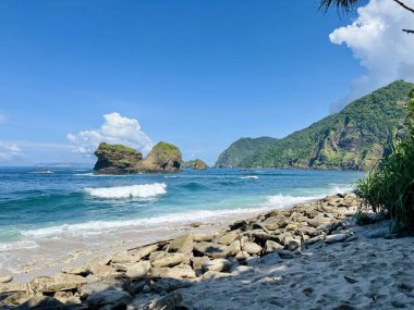 Beyaz kumlu tropik sahil manzarası, kıyı kayaları, ve mavi gökyüzünün altındaki yemyeşil tepeler.