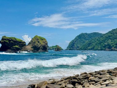 Çarpışan dalgalarla tropik deniz manzarası, yeşil tepeler ve bulutlu mavi gökyüzü.