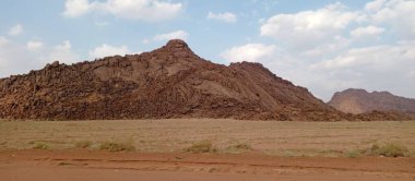 Parlak mavi gökyüzünün altında, dağınık bulutların altında, çöl manzarasında çarpıcı kayalık bir dağ oluşumu..
