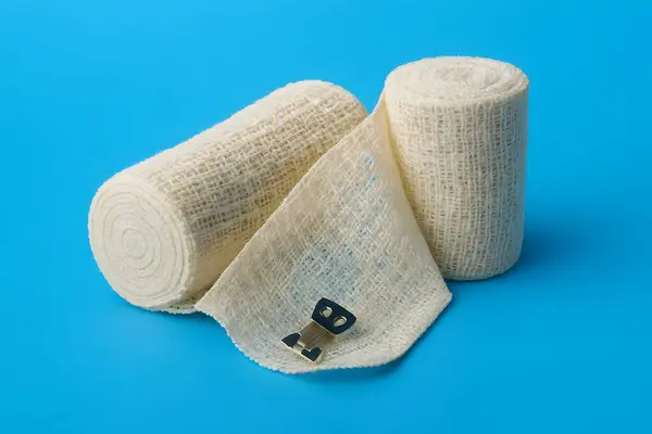 Parlak mavi arkaplanda iki beyaz sargı bezi rulosu, biri kısmen açılmış metalle sarılmış steril yara bakımı ve solunabilir pansuman malzemesi..