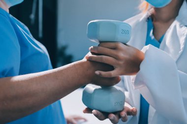 Evde oturma odasında terapi ve rehabilitasyon görürken, bakıcı ya da fizyoterapist yaşlılara yardım ederken, son sınıf öğrencisi olan Asyalı bir kadın ya da hemşire kol egzersizi için halteri kaldırıyor..