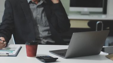 Takım elbiseli ve gözlüklü genç işadamı ofiste dizüstü bilgisayarla çalışıyor.