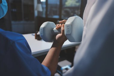 Evde oturma odasında terapi ve rehabilitasyon görürken, bakıcı ya da fizyoterapist yaşlılara yardım ederken, son sınıf öğrencisi olan Asyalı bir kadın ya da hemşire kol egzersizi için halteri kaldırıyor..