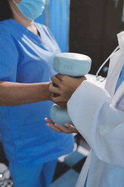 Evde oturma odasında terapi ve rehabilitasyon görürken, bakıcı ya da fizyoterapist yaşlılara yardım ederken, son sınıf öğrencisi olan Asyalı bir kadın ya da hemşire kol egzersizi için halteri kaldırıyor..