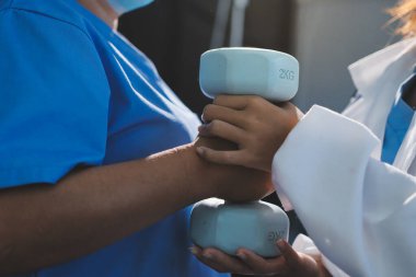 Evde oturma odasında terapi ve rehabilitasyon görürken, bakıcı ya da fizyoterapist yaşlılara yardım ederken, son sınıf öğrencisi olan Asyalı bir kadın ya da hemşire kol egzersizi için halteri kaldırıyor..