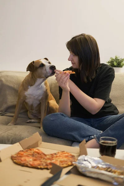 A woman eating pizza at home and sharing a bite with her dog. Ready delivered meals, eating at home with dog on the couch