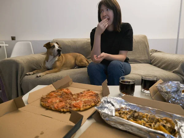 家でピザを食べて犬を見ている面白い女性 ソファで犬と一緒に家で食事をする準備ができている ストック写真