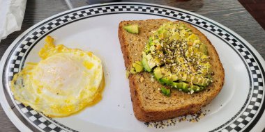 Simitle kaplı bir dilim avokado tostu ve kareli kenarlı mükemmel pişmiş yumurta içeren lezzetli bir kahvaltı tabağı..