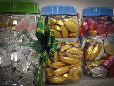 Her biri bir mağaza rafında dekoratif bir kurdeleyle süslenmiş gümüş, altın ve pembe ambalajlar içeren paketlenmiş çikolata şeker kavanozlarının görsel çekiciliği..