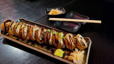 Taze soğanla süslenmiş, lezzetli soslu, kıtır kıtır bir suşi rulosunun yakın plan görüntüsü. Yakınlarda, yemek çubukları ilave zencefil ve wasabi turşusunun yanında dinleniyor..