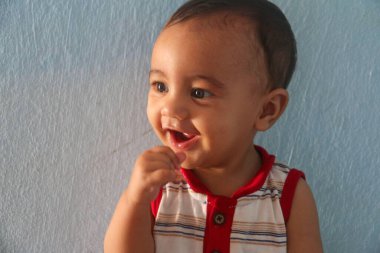 Güzel bir bebek mutlu bir şekilde gülümser, parlak bebek dişlerini gösterir. Kırmızı ve beyaz çizgili bir gömlek giyerek, gözlerinde bir pırıltıyla kameraya bakar, çocukluk ve masumiyetin özünü yakalar. Yumuşak mavi arka plan onun mutlu ifadesini arttırır.