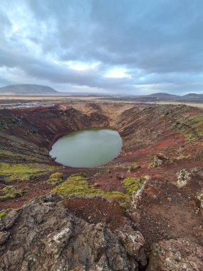 A stunning view of a volcanic crater lake shrouded in a cloudy sky. The landscape features exposed rocky terrain with patches of green moss, set against distant rolling hills. clipart