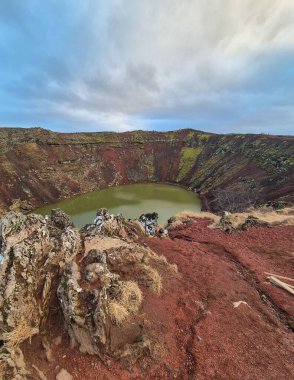  a green lake in a volcanic crater shrouded in a lightly cloudy sky. The rugged landscape features exposed rocky terrain with patches of green moss, set against rolling hills in the distance clipart