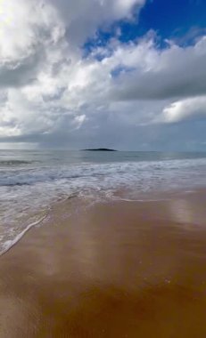 Okyanusun yumuşak dalgalarını yakalayan sakin sahil manzarası ufuk çizgisinde görünen bir adayla, parçalı bulutlu bir gökyüzünün altında kumlu bir sahile vuruyor.