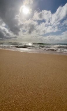 Dalgaların kırılma görüntüsü düşük, bulutlar denizin üzerine gölgeler yansıtıyor, sakin ama güçlü bir atmosfer yaratıyor. Güneş bulutların arasından süzülüyor ışıldayan dalgaları aydınlatıyor.