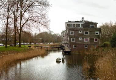 10-03-2024 - Amsterdam, Hollanda - eğimli cepheli yenilikçi ahşap bina sakin bir su yolu üzerinde duruyor, bir yaya köprüsü ile bağlanıyor, modern tasarımı kentsel bir çevrede doğal çevreyle harmanlıyor. Hollanda, Amsterdam 'da yakalandı.,