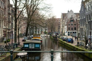 10-03-2024 - Amsterdam, Hollanda - Amsterdam kanalı büyüleyici yüzen tekneler ve geleneksel Hollanda binalarıyla doludur. Bisikletler kaldırımlı caddeler boyunca dinlenirken yayalar sakin suların yanında gezerken şehir manzarasını yansıtıyorlar. Yakaladım!