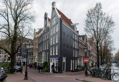 10-03-2024-Amsterdam, Hollanda, zarif dar Amsterdam kanal evi koyu siyah cepheli, keskin gaçalı çatısı ve tarihi mimarisi var. Kanallar boyunca yürüyen bisikletlerle, ağaçlarla ve yayalarla çevrili büyüleyici bir sokak köşesi. A 'da Yakalandı