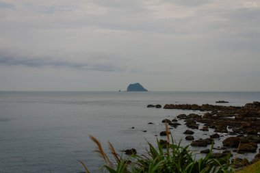 Sakin kıyı sahneleri sakin sulara uzanan kayalık bir kıyı şeridini, ufukta bulutlu bir gökyüzünün altındaki uzak bir adanın barışçıl bir deniz manzarası yarattığını gösteriyor. Tayvan, Asya 'da yakalandı..