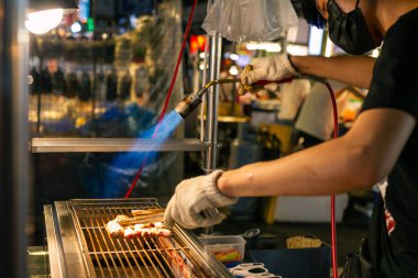 14-08-2023 - Taipei, Tayvan: Canlı gece pazarında aç ziyaretçilere lezzetli sokak yemekleri hazırlamak için ızgara et şişleri yakmak ve tatlandırmak için kaynak makinesini ustalıkla kullanan yetenekli bir sokak satıcısı. Tayvan, Asya 'da yakalanmış..