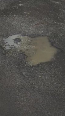 Karanlık bir asfalt yolda bir su birikintisi, yağmur damlaları su yüzeyinde dalgalanmalar ve daireler yaratır..