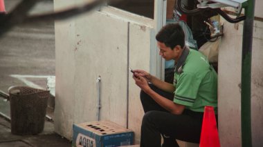 Yeşil üniformalı genç bir adam telefonuna dalmış beton bir duvara yaslanmış oturuyor. Sahne açık havada, muhtemelen şehir ortamında, yakınlarda bir kutu su şişesi var..