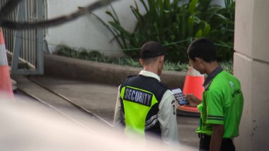 Üniformalı iki güvenlik görevlisi akıllı telefondaki kamera görüntülerini inceliyor. Sahne açık havada, arka planda trafik konileri ve yeşillik var..